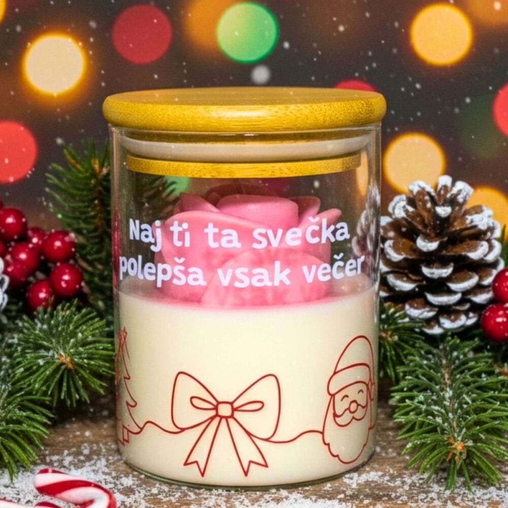 Božična dišeča sveča z napisom "Naj ti ta svečka polepša vsak večer", z roza vrtnico, postavljena v aranžma z jelko, storži in božičnimi lučkami v ozadju