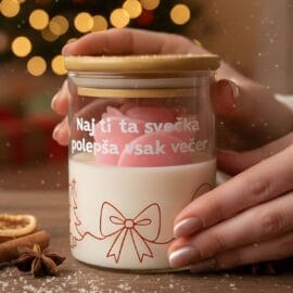 Roke držijo dišečo svečo darilo z napisom "Naj ti ta svečka polepša vsak večer", z roza vrtnico in božičnim motivom, na leseni mizi z začimbami in zamegljenim božičnim drevescem v ozadju.