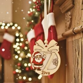 Leseni personaliziran božični okrasek z motivom Božička, izrezljano črko 'Ž' in imenom 'Živa', obešen na kaminu s praznično dekoracijo v ozadju.