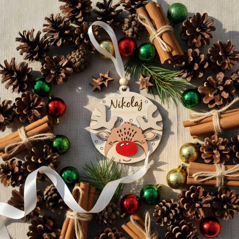 Leseni personaliziran okrasek Jelenček z rdečim noskom, z imenom 'Nikolaj', položen v krog naravne dekoracije: storži, cimet, rdeče in zelene bunkice.