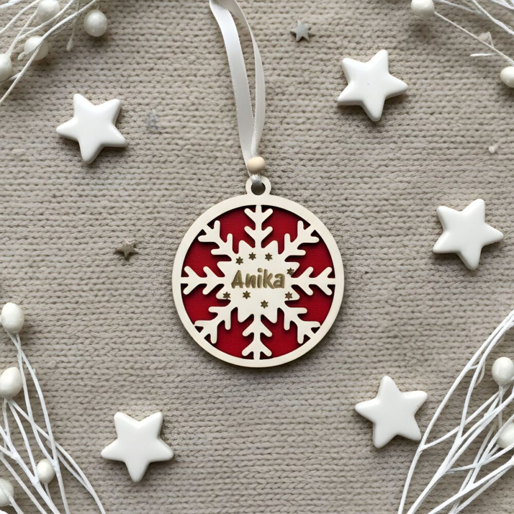 Dvoslojni personaliziran okrasek Snežinka z rdečo podlago in imenom 'Anika', položen na toplem pletenem ozadju, obkrožen z belimi zvezdicami in okrasnimi vejicami.