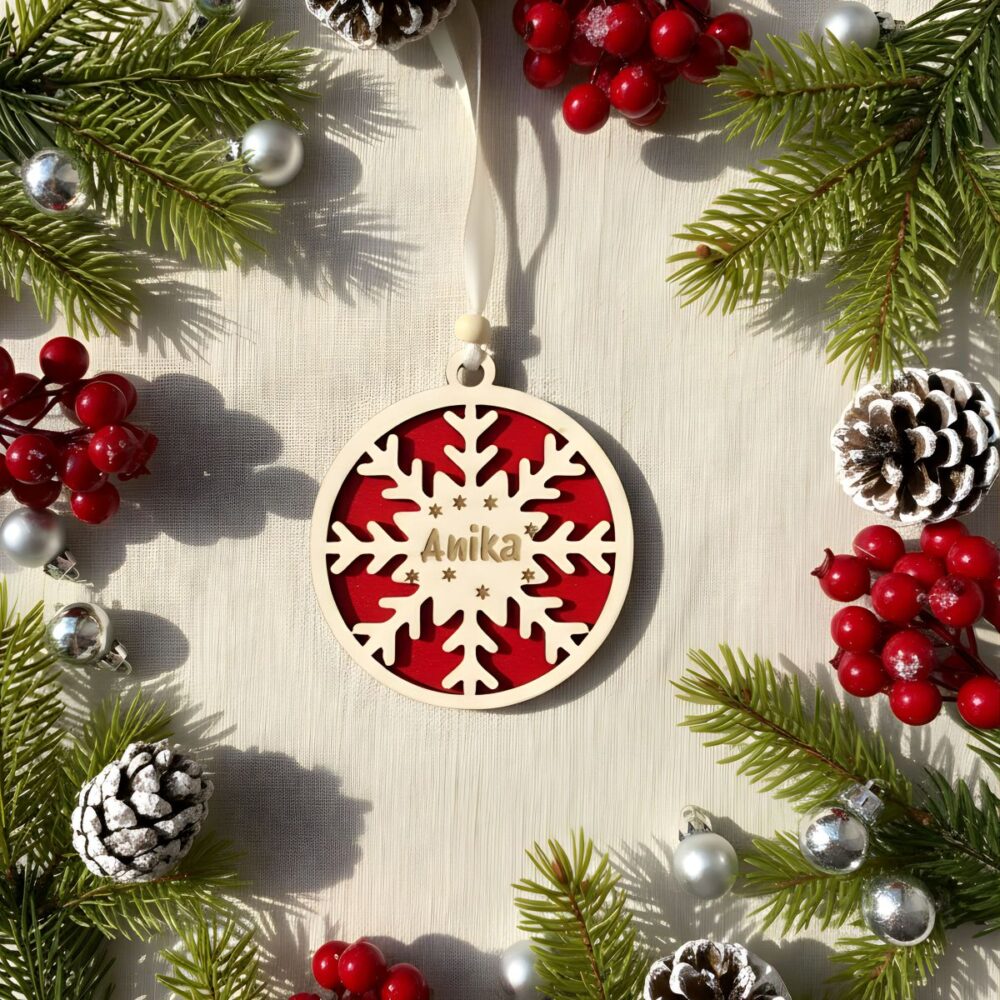 Božični okrasek iz lesa s snežinko in vgraviranim imenom Anika, rdeče ozadje, v okviru smrekovih vej, storžev in rdečih jagod.