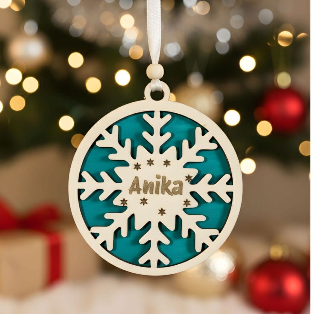 Dvoslojni personaliziran okrasek Snežinka z vgraviranim imenom 'Anika' in zeleno podlago, obešen pred zamegljenim božičnim drevescem z rdečimi in zlatimi okraski.