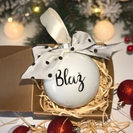 Personaliziran steklen božični okrasek z imenom "Blaž" in črnim napisom, okrašen z belo pentljo s črnimi pikami, v darilni škatli, na prazničnem božičnem ozadju z lučkami.