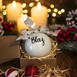 Personaliziran steklen božični okrasek z imenom "Blaž" in črnim napisom, okrašen z belo pentljo s črnimi pikami, v darilni škatli. Ozadje prikazuje prižgane sveče, praznične lučke in božično dekoracijo.