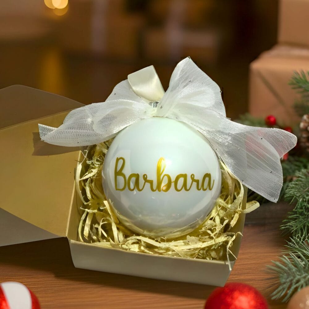 Personaliziran steklen božični okrasek z imenom "Barbara" in sijočim zlatim napisom, okrašen z veliko belo pentljo, postavljen v odprto darilno škatlo na leseni podlagi z zamegljenim božičnim ozadjem.