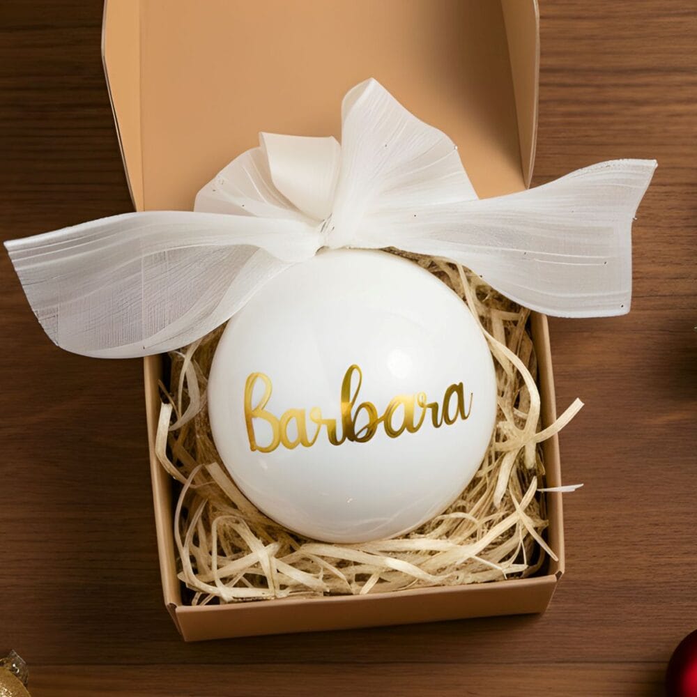 Beli steklen božični okrasek z bleščečim zlatim napisom "Barbara" v pisavi, ki spominja na kaligrafijo. Okrasek je okrašen z veliko belo, prosojno pentljo in postavljen v rjavo darilno škatlo na leseni mizi.