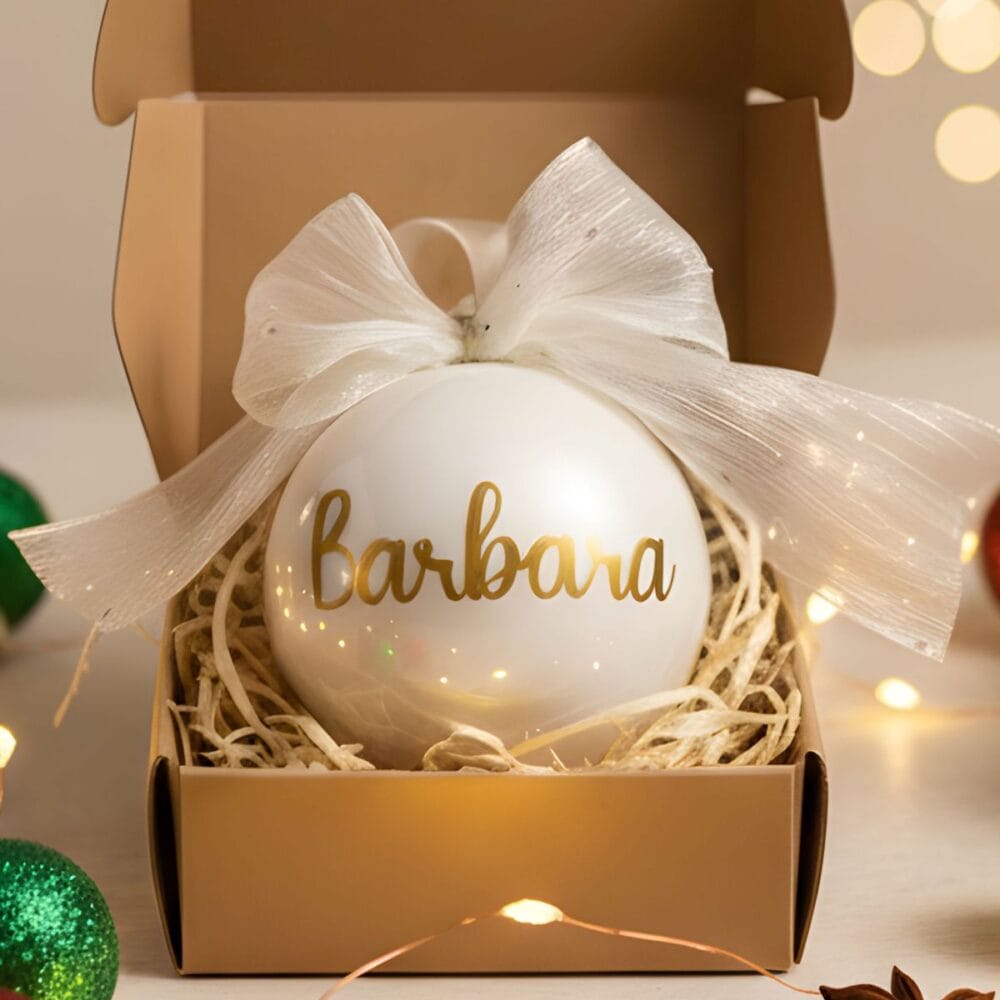 Personaliziran beli steklen božični okrasek z imenom "Barbara" in zlatim napisom, postavljen v darilno škatlo in obdan s sijočimi božičnimi lučkami ter zelenimi in rdečimi okraski v ozadju.