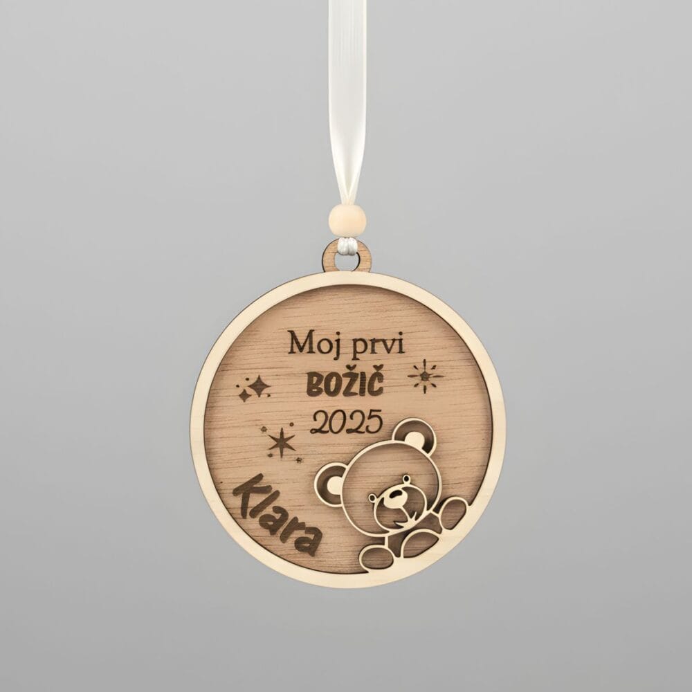 Leseni personaliziran okrasek 'Moj Prvi BOŽIČ' z imenom 'Klara' in letnico '2025' ter motivom medvedka, obešen na belem traku pred enobarvnim sivim studijskim ozadjem.