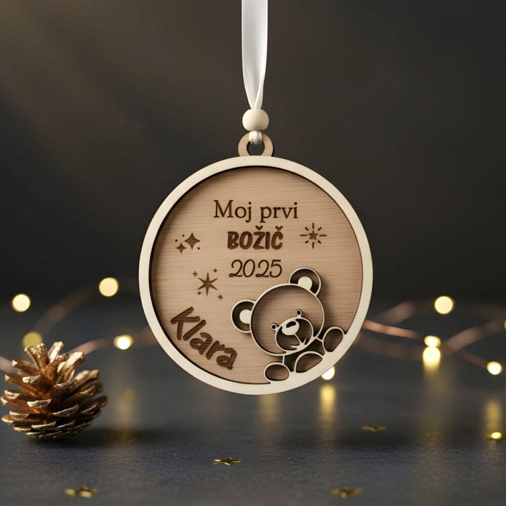 Leseni personaliziran okrasek 'Moj Prvi BOŽIČ' z imenom 'Klara' in letnico '2025' ter motivom medvedka, obešen pred temnim ozadjem z lučkami in zlatim storžem.