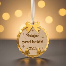 Elegantni personaliziran okrasek z zlatimi detajli za 'Matejev prvi božič 2025', obešen pred zamegljenim zlatim božičnim ozadjem.