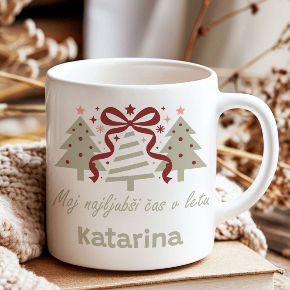 Bela keramična skodelica z božičnim motivom, ki prikazuje tri stilizirane jelke v sivih in rdečih tonih. Na skodelici je napis 'Moj najljubši čas v letu' in personalizirano ime 'Katarina'. Skodelica je postavljena na kup knjig, obdana z mehko pleteno odejo, kar ustvarja udoben zimski prizor.