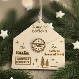 Leseni personaliziran okrasek v obliki kuverte z napisom 'Pismo za Božička' in 'Od: Marka', z žigom 'Ekspresna pošta', položen na smrekovih vejicah z belimi zvezdicami.
