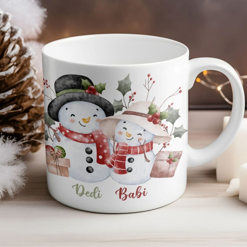 Božična skodelica za dedija in babico z ilustracijo dveh snežakov, oblečenih v rdeče šale in kape, ter napisoma "Dedi Babi", postavljena v zimsko dekoracijo.