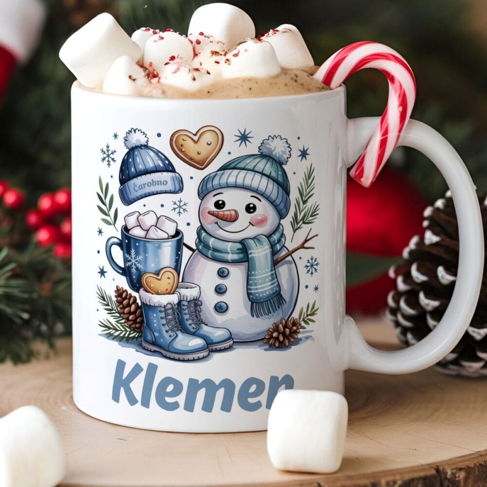 Bela keramična skodelica z zimsko ilustracijo snežaka v modrem šalu in kapi. Na skodelici je ime Klemen. Skodelica je polna vroče čokolade, marshmallowov in rdeče-bele sladkorne palice, postavljena na leseno podlago