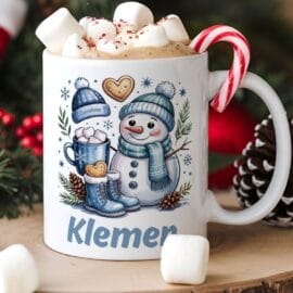 Bela keramična skodelica z zimsko ilustracijo snežaka v modrem šalu in kapi. Na skodelici je ime Klemen. Skodelica je polna vroče čokolade, marshmallowov in rdeče-bele sladkorne palice, postavljena na leseno podlago