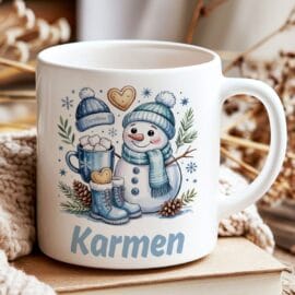 Bela keramična skodelica z motivom nasmejanega snežaka v modrih oblačilih in personaliziranim imenom Karmen. Skodelica stoji na kupčku knjig ob topli pleteni odeji in suhi zimski dekoraciji
