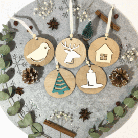 Set petih lesenih božičnih okraskov na sivi podlagi z izrezanimi snežinkami. Okraski (ptica, jelen, hiška, smrečica, svečka) so obdani z vejicami evkaliptusa, cimetovimi palčkami, zvezdastim janežem in borovimi storži.