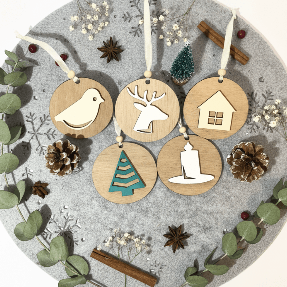 Set petih lesenih božičnih okraskov na sivi podlagi z izrezanimi snežinkami. Okraski (ptica, jelen, hiška, smrečica, svečka) so obdani z vejicami evkaliptusa, cimetovimi palčkami, zvezdastim janežem in borovimi storži.