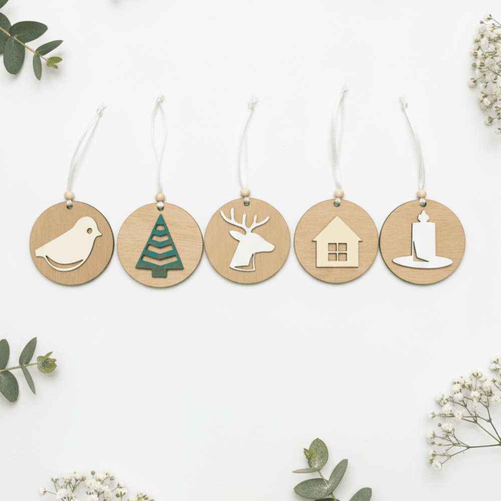 Set petih lesenih božičnih okraskov v ravni vrsti na beli minimalistični podlagi. Okraski (ptica, smrečica, jelen, hiška, svečka) so dekorirani z vejicami evkaliptusa in cvetovi gipsofilke