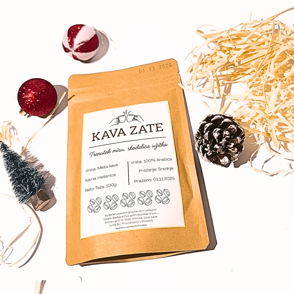 Paket mlete kave "Kava Zate" na beli podlagi, obdan z božičnimi okraski. Vidni so rdeči okraski, majhno umetno božično drevesce, storž in slama. Besedilo na paketu vključuje "Kava Zate", "Vrsta: Mleta kava", "Kavna mešanica", "Vrsta: 100% Arabica", "Praženje: Srednje", "Neto Teža: 100g", "Praženo: 01.11.2025" in podatke o proizvajalcu.