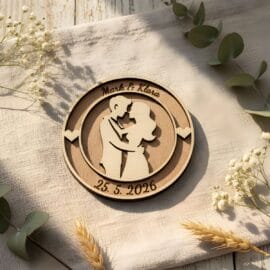Personaliziran okrogel lesen magnet z vgravirano silhueto objemajočega se para, imenoma Mark & Klara ter datumom, fotografiran v topli naravni svetlobi na bež tekstilni podlagi z dekoracijo evkaliptusa in suhega cvetja.