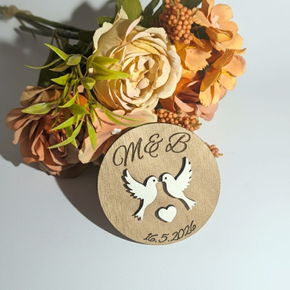 Personaliziran poročni magnet z vgraviranima začetnicama M & B, datumom poroke in motivom golobčka ljubezni, položen ob šopek suhega cvetja v mareličnih in oranžnih odtenkih na beli podlagi.