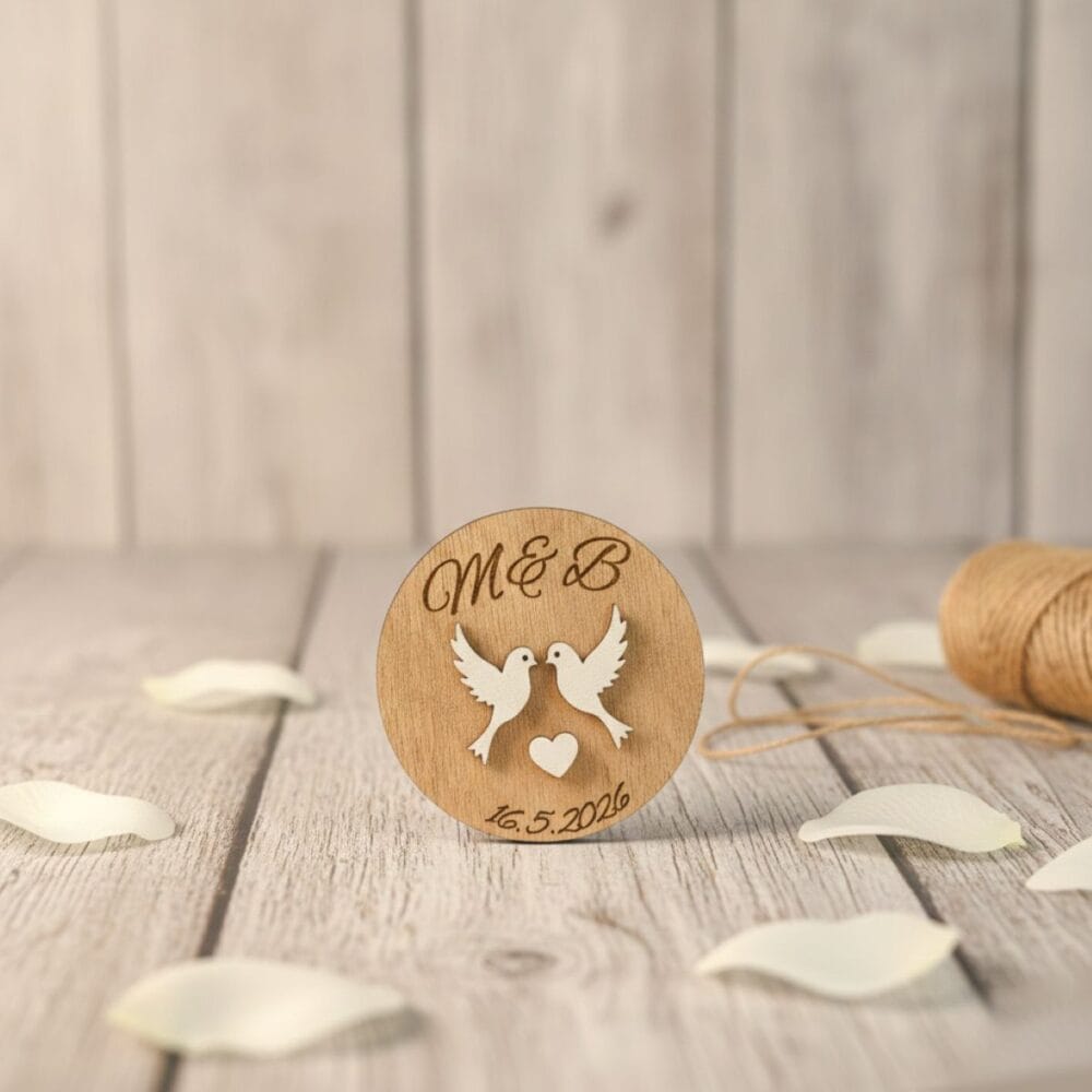 Personaliziran leseni poročni magnet z vgraviranimi začetnicami M & B in datumom, motivom dveh belih golobčkov ter srcem, postavljen na sivo leseno površino s klobčičem vrvice v ozadju.