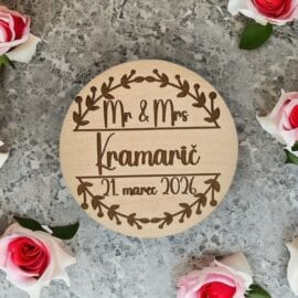 Personaliziran okrogel leseni poročni magnet z graviranim napisom Mr & Mrs Kramarič in datumom poroke, obdan z nežnimi rožami na sivi kamniti podlagi kot unikatna poročna zahvala.