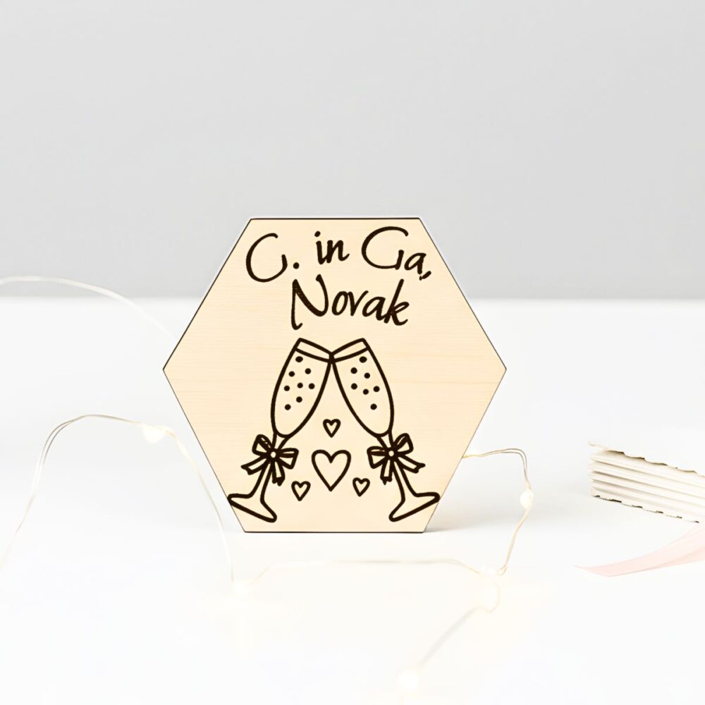Personaliziran šesterokoten lesen poročni naprsnik z vgraviranima imenoma G. in Ga. Novak, motivom dveh kozarcev šampanjca in srčki, postavljen v minimalističnem ambientu z elegantnimi lučkami v ozadju.