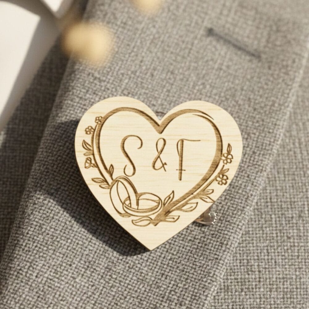 Personaliziran lesen poročni naprsnik v obliki srca z vgraviranima prepletenima prstanoma in začetnicama S & T, pritrjen na rever sive moške poročne obleke.