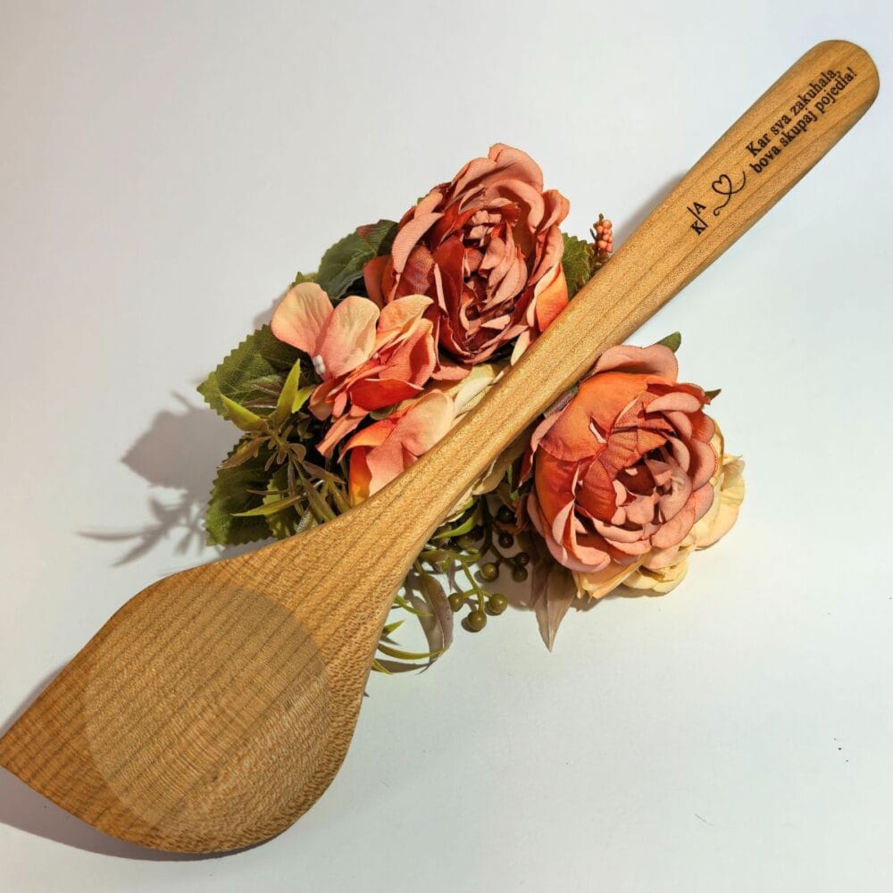 Personalizirana kuhalnica iz češnjevega lesa z vgraviranim napisom 'Kar sva zakuhala, bova skupaj pojedla' in začetnicami, položena na dekorativno cvetje na beli podlagi.