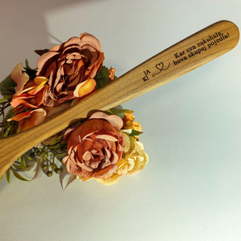 Bližnji posnetek ročaja kuhalnice iz češnjevega lesa s podrobno gravuro 'Kar sva zakuhala, bova skupaj pojedla!', začetnicama in srčkom, na estetskem rožnatem ozadju s cvetnimi lističi.