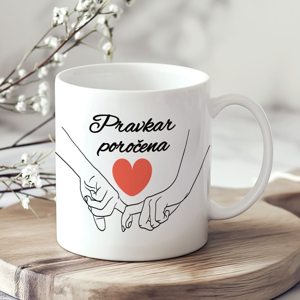 Bela keramična skodelica z napisom "Pravkar poročena" v črni pisavi, pod katero je ilustracija dveh rok, ki se držita za mezinca, in rdeče srce. Skodelica stoji na lesenem podstavku ob nežnem cvetju.