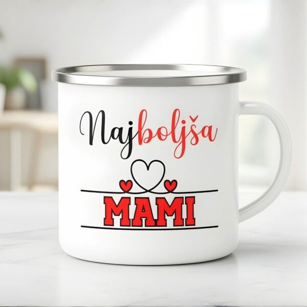 Bel emajliran lonček za mamo s srebrnim robom, ki prikazuje minimalističen napis 'Najboljša MAMI' z rdečimi srčki na svetlem, zamegljenem ozadju kuhinje.