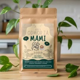 Vrečka mlete kave 'Odmor za mami' s slovenskim napisom in ilustracijo mame z dojenčkom, postavljena na leseno mizo ob zeleni sobni rastlini.
