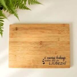 Detajl lesene rezalne deske z napisom za mamo, postavljene zraven zelene praproti v svetli, moderni kuhinji.