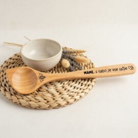 Lesena kuhalnica za mami s čebelico in vgraviranim napisom "MAMI, s tabo je vse lažje", položena na pleten podstavek ob beli glineni skledici in suhem cvetju sivke.