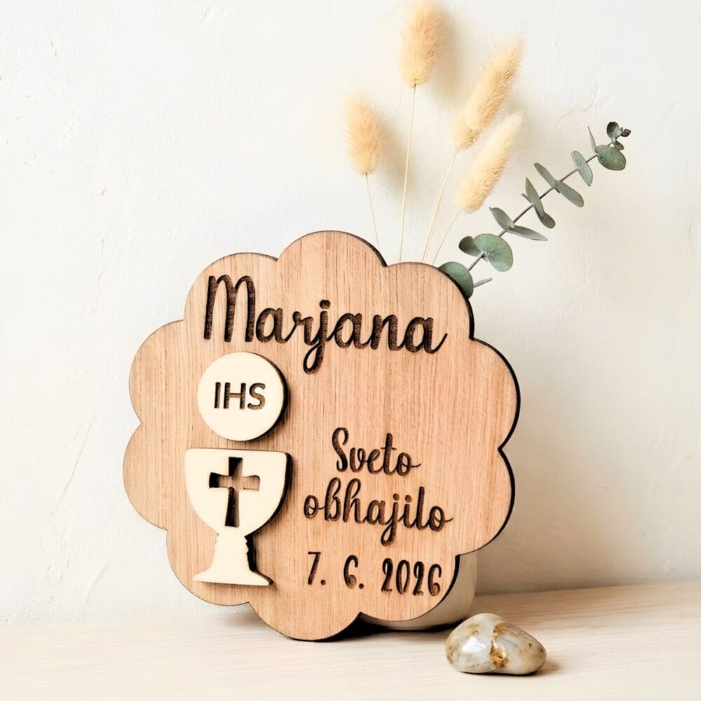 Leseni magnet za prvo sveto obhajilo v obliki oblačka z reliefnim kelihom, hostijo z napisom IHS, vgraviranim imenom Marjana in datumom, postavljen ob dekorativno vazo s suhim cvetjem.