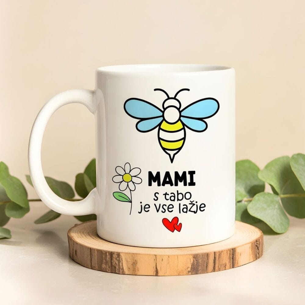 Bela keramična skodelica z ilustracijo barvne čebelice, marjetice in napisa "MAMI s tabo je vse lažje", postavljena na lesen podstavek z zelenjem v ozadju.