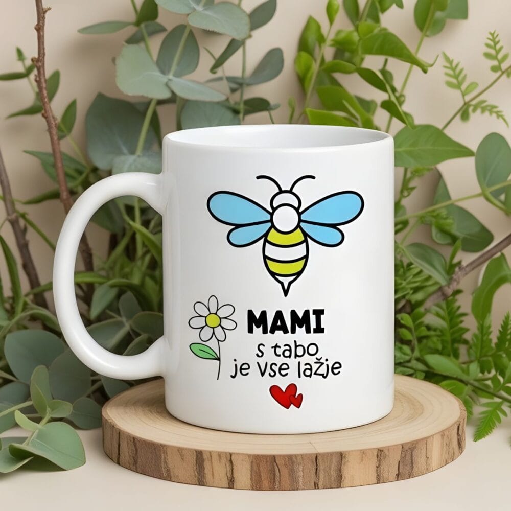 Skodelica za mami s čebelico in napisom "MAMI, s tabo je vse lažje", postavljena na ličen lesen podstavek, obdana z bogatimi vejami evkaliptusa in praproti v ozadju.