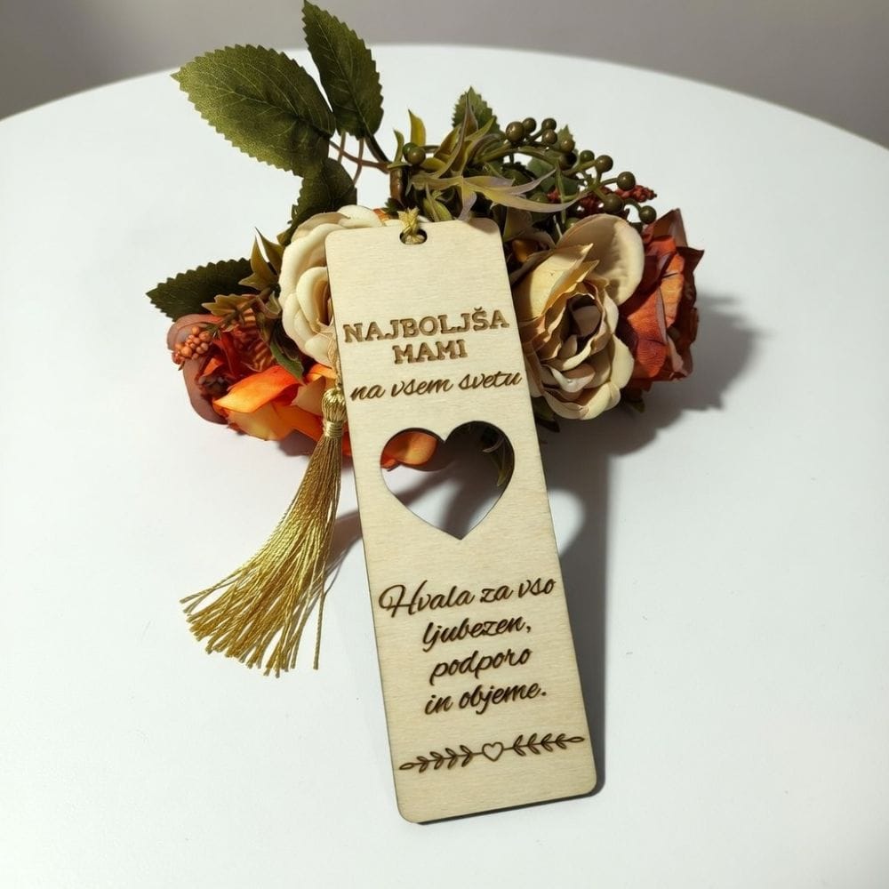 Leseno knjižno kazalo z vgraviranim napisom "NAJBOLJŠA MAMI na vsem svetu" in izrezanim srčkom, okrašeno z zlatim čopičem, položeno ob šopek umetnih cvetlic.