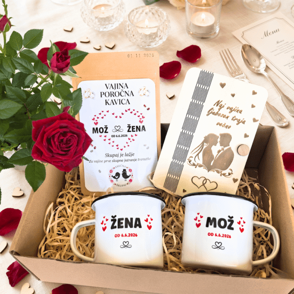 Darilni paket za mladoporočenca na poročni mizi, ki vsebuje personalizirani emajlirani skodelici Mož in Žena, poročno kavo in gravirano leseno darilo, obkroženo z rdečimi vrtnicami in poročnim dekorjem.
