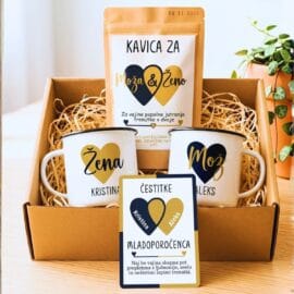 Darilni set za mladoporočenca v kartonasti škatli z naravnim polnilom. Vsebuje dve personalizirani emajlirani skodelici z napisoma "Žena Kristina" in "Mož Aleks" ter paket kave z etiketo "Kavica za moža & ženo". V ospredju je poročna čestitka z napisom "Čestitke mladoporočenca" in osebnim posvetilom.